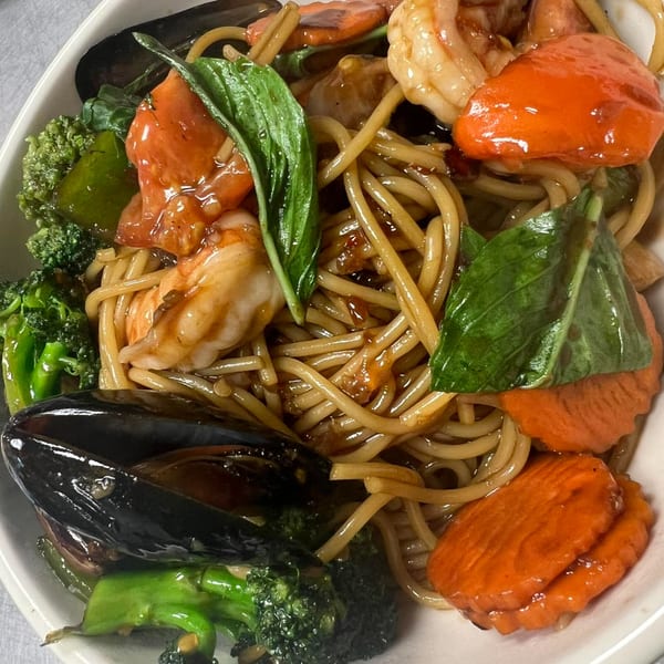 Thai noodles with shrimp, mussels, broccoli, carrots, and basil in a white bowl.