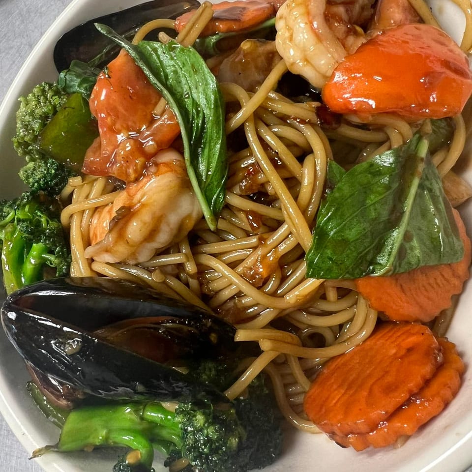 Thai noodles with shrimp, mussels, broccoli, carrots, and basil in a white bowl.