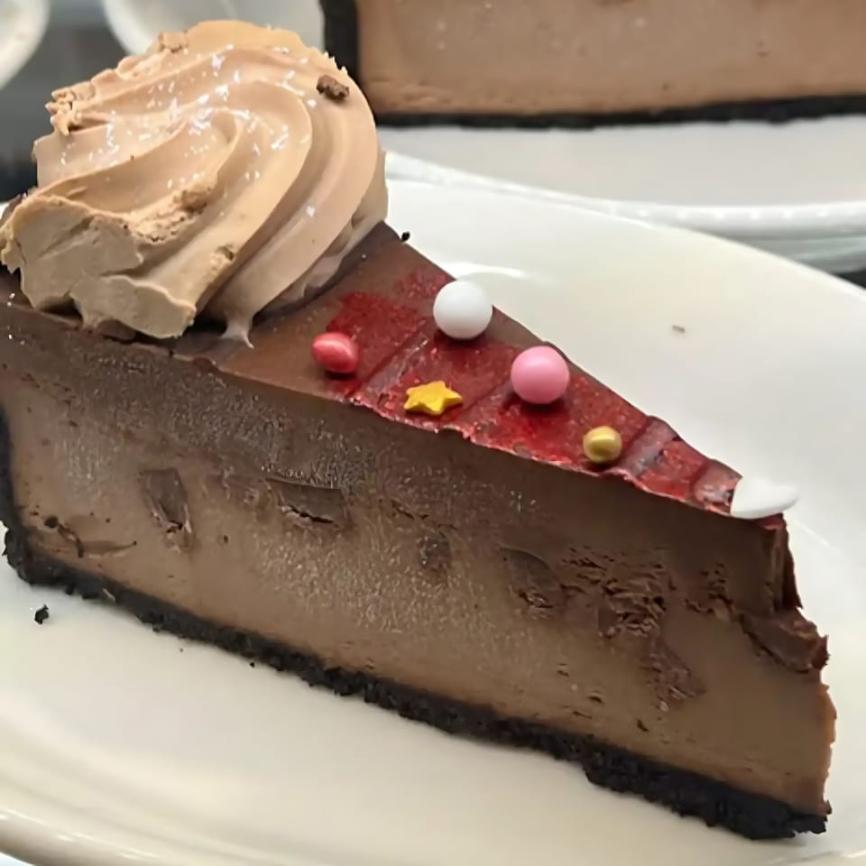 A slice of chocolate cheesecake with whipped cream and colorful sprinkles on a white plate.
