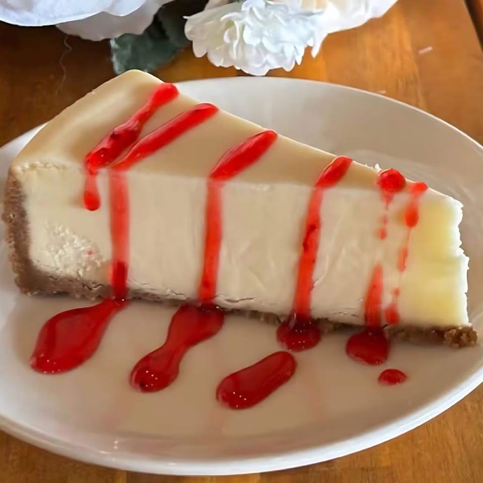 Slice of cheesecake with a graham cracker crust, drizzled with red raspberry sauce on a white plate.