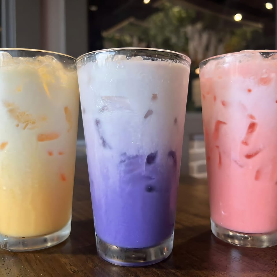 Three vibrant Thai iced drinks: yellow, purple, and pink, with ice, on a wooden table.