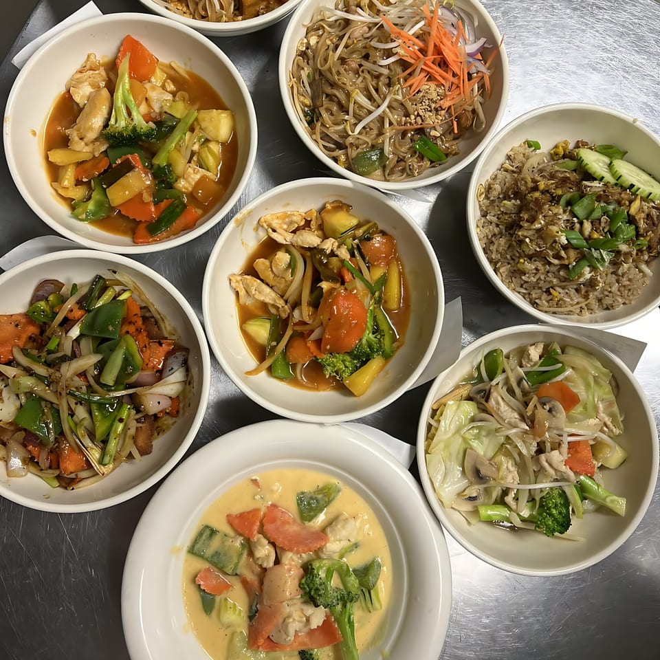 An overhead view of seven bowls of various Thai dishes, including curries, noodles, and stir-fries.