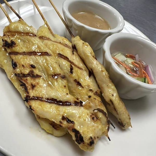 Grilled chicken satay skewers with peanut sauce and pickled vegetables on a white plate.