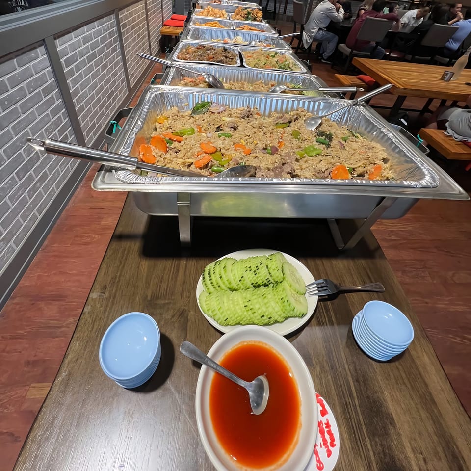 A Thai buffet line with various dishes, including fried rice, and a table setting with sauce and cucumbers.