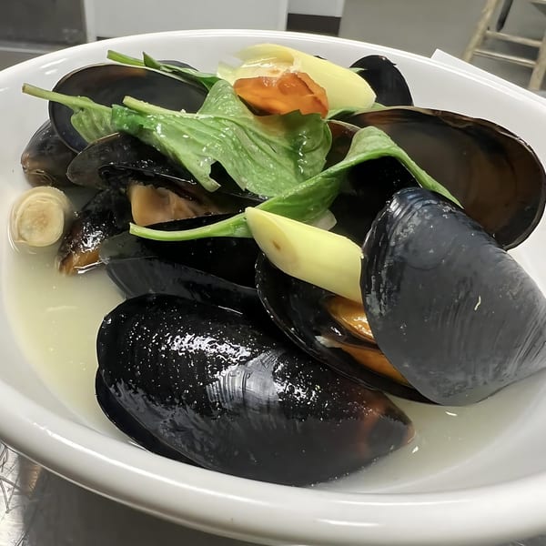 Steamed mussels in a white bowl with broth, garnished with green herbs and sliced vegetables.